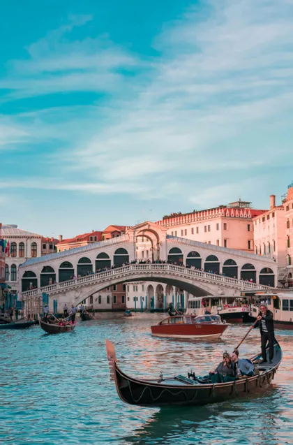 A gondolier rows a gondola with passengers on a busy canal, with a large, arched stone bridge in the background under a blue sky.