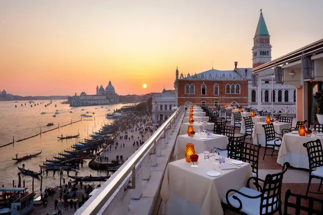 Set tables on a rooftop restaurant overlook a historic city's waterfront with boats and gondolas at sunset.