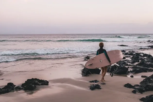 Ein Surfer in einem Neoprenanzug trägt ein pinkes Surfbrett und geht in der Abenddämmerung über einen sandigen und felsigen Strand zum Meer.