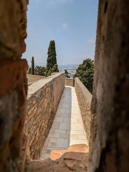 Una vista attraverso un'apertura in pietra di uno stretto camminamento pavimentato su una mura della fortezza, con cipressi e un paesaggio urbano in lontananza.