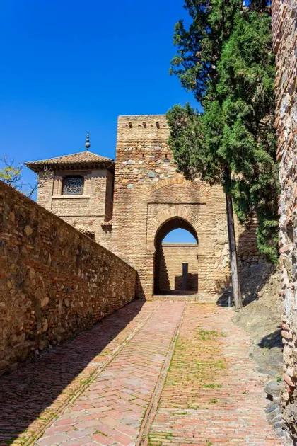 Un camino empedrado conduce a un arco de entrada en una fortaleza de piedra histórica bajo un cielo azul brillante.