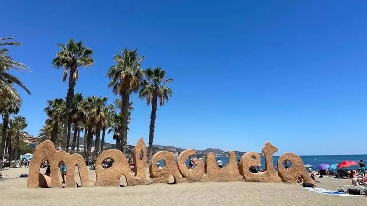 A large sand sculpture spelling 'Malagueta' on a sunny beach with tall palm trees and the ocean in the background.
