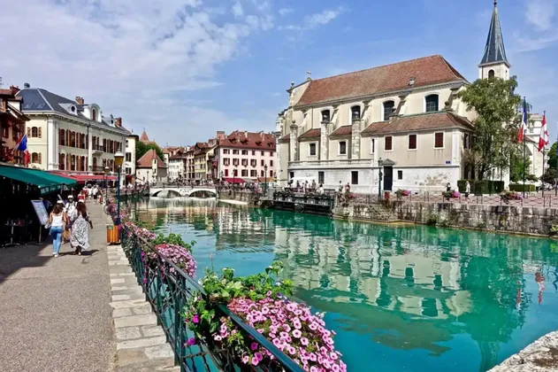 Des personnes se promènent le long d'une promenade fleurie, bordant un canal turquoise, avec des bâtiments historiques et une église se reflétant dans l'eau.