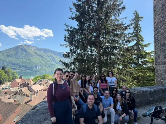 A WeRoad group trip poses for a photo on a stone wall, with a lakeside town and green mountains in the background.