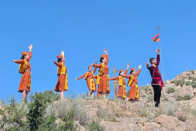 Eine Gruppe von Personen in traditionellen orange-gelben Kostümen vollführt einen synchronisierten Tanz auf einem felsigen Berghang unter strahlend blauem Himmel, neben einer armenischen Flagge.
