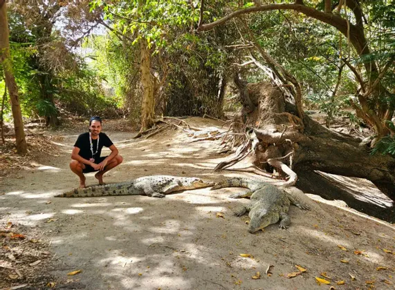 Ein lächelnder Mann hockt barfuß auf einem Sandweg neben drei großen Krokodilen, mit einem üppig grünen Wald im Hintergrund.