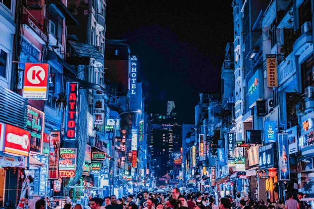 Eine belebte Stadtstraße bei Nacht, auf der Menschen unter hohen Gebäuden mit leuchtenden blauen und roten Neonschildern entlanglaufen.