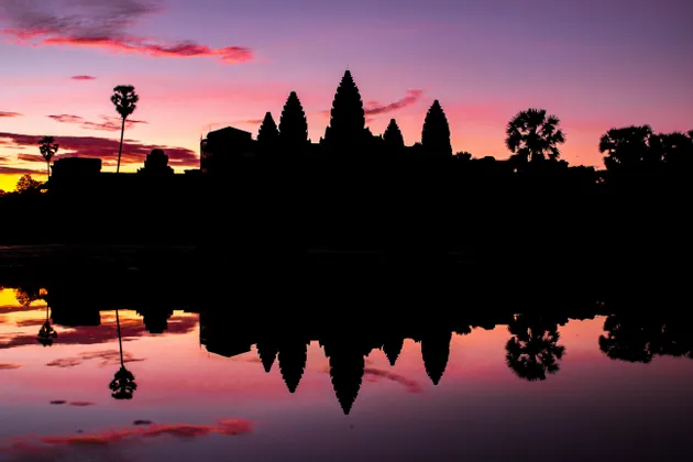 Silhouette di un complesso di templi e palme riflesse in acqua calma durante un tramonto rosa e viola.