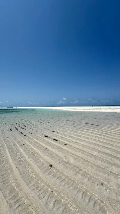 Ondulazioni di sabbia in acqua bassa e trasparente su una spiaggia di sabbia bianca sotto un vasto cielo azzurro e senza nuvole.