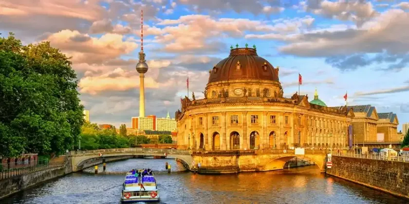 Una barca turistica naviga su un fiume cittadino, passando davanti a un palazzo dalla cupola ornata, con un'alta torre della televisione in lontananza al tramonto.