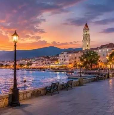 Passeggiata pavimentata sul lungomare con lampione illuminato e vista su una città costiera con campanile al tramonto.