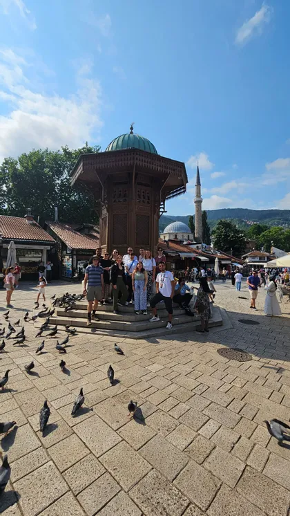 Eine WeRoad-Gruppe posiert an einem sonnigen Tag auf einem gepflasterten Platz mit einem verzierten Holzbrunnen und vielen Tauben.