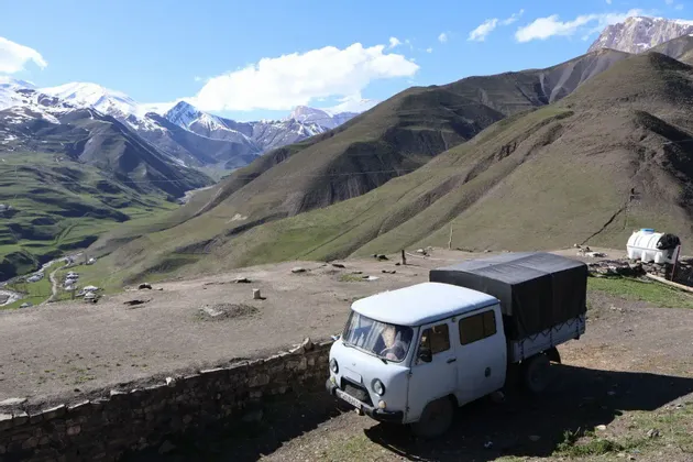 Ein Van parkt auf einem Feldweg an einem Berghang, mit Blick auf ein grünes Tal und entfernte schneebedeckte Berge.