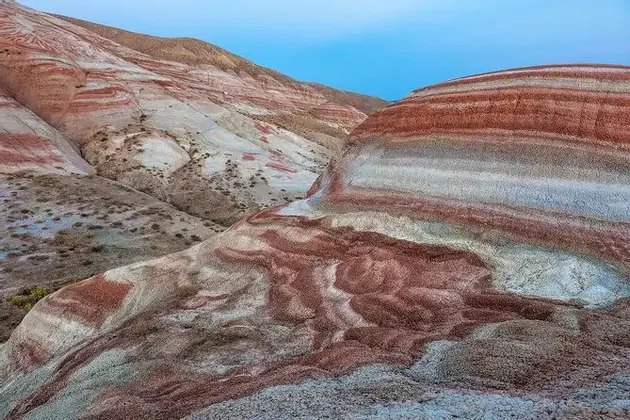 Schichten aus rotem, weißem und grauem Sediment bilden sanfte, gestreifte Hügel unter einem hellblauen Himmel.