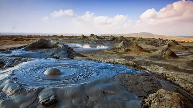 Ein blubbernder Schlammvulkan erzeugt konzentrische Wellen in einer ariden Landschaft mit zahlreichen kleineren Schlammhügeln unter bewölktem Himmel.