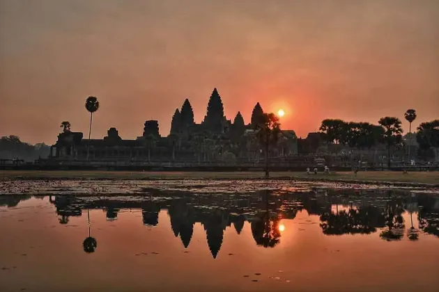 Bei orangefarbenem Sonnenaufgang spiegelt sich die Silhouette eines großen, antiken Tempels mit mehreren Türmen in einem stillen Gewässer.