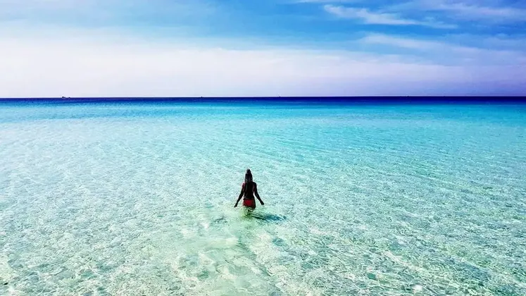 Eine Frau mit dem Rücken zur Kamera geht in das seichte, klare türkisfarbene Meer unter einem strahlend blauen Himmel.