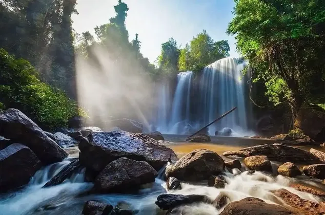 Ein breiter Wasserfall stürzt in einen felsigen Fluss in einem dichten Wald, während Sonnenlicht durch die Bäume und den Nebel strömt.
