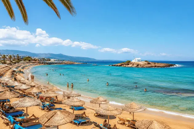 Una veduta dall'alto di una spiaggia sabbiosa affollata con ombrelloni di paglia, persone che nuotano nell'acqua turchese e una piccola isola con una cappella.