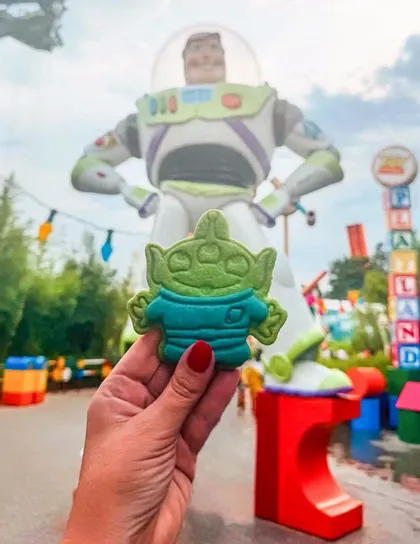 Una mano con smalto rosso tiene un biscotto verde a forma di alieno davanti a una grande statua sfocata di Buzz Lightyear.