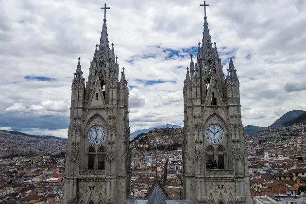 Dos torres de reloj de catedral de estilo gótico dominan una ciudad extensa con colinas al fondo bajo un cielo nublado.