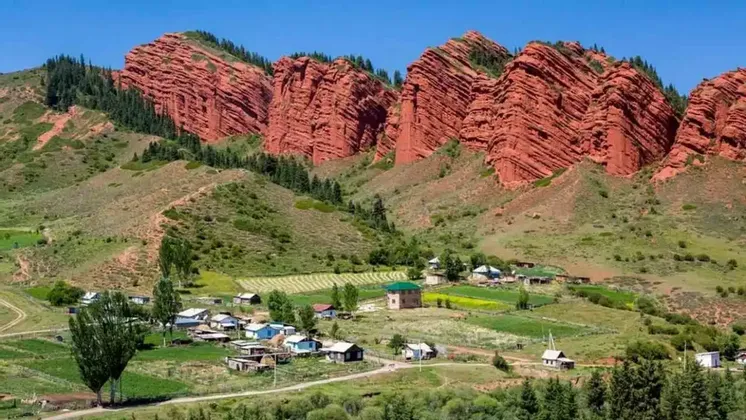 Un piccolo villaggio con campi si trova in una lussureggiante valle verde ai piedi di imponenti montagne di roccia rossa stratificata sotto un cielo sereno.