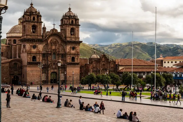 Una ornamentada catedral de piedra con campanarios gemelos domina una concurrida plaza pavimentada llena de gente, con montañas verdes de fondo.