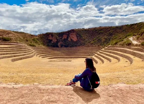 Une femme avec un sac à dos est assise sur un rebord de terre surplombant un grand site archéologique circulaire en terrasses sous un ciel nuageux.