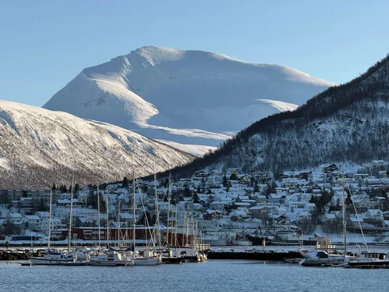 Sailboats are docked in a harbor in front of a snowy town nestled at the base of large, snow-covered mountains.