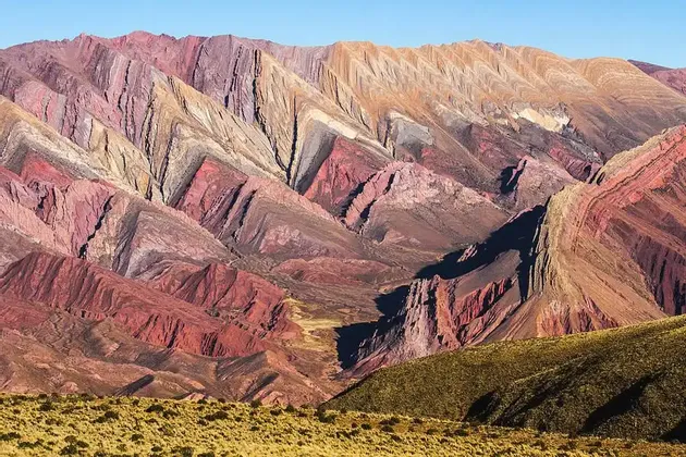 Una catena montuosa mostra formazioni rocciose colorate e stratificate dalle tonalità di rosso, arancione e viola, sotto un cielo azzurro e limpido.