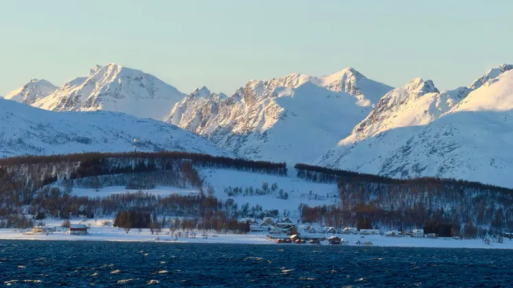 A small village on a snowy coast with blue water in the foreground and a large, sunlit mountain range behind.