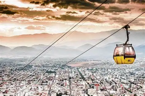 Una funivia gialla si muove su cavi ad alta quota sopra una città, con una catena montuosa visibile in lontananza sotto un cielo drammatico.