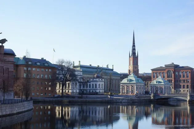 Uno scorcio urbano con edifici storici e un campanile di chiesa che si riflettono nell'acqua placida di un canale in una giornata di sole.