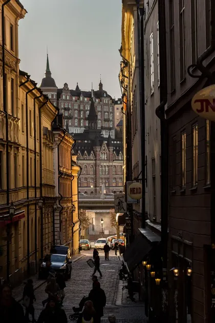 Una vista su una stretta via acciottolata, fiancheggiata da alti edifici storici, con persone che passeggiano alla luce della sera.