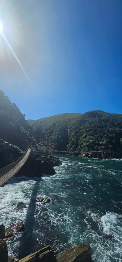 Eine Hängebrücke spannt sich an einem hellen, sonnigen Tag über tosende Küstengewässer zu dichten, grünen Hügeln hin.