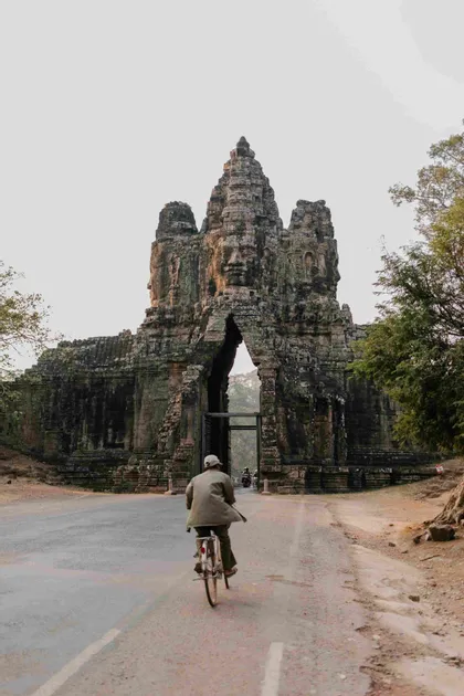 Una persona de espaldas pedalea en bicicleta por una carretera hacia una monumental y antigua puerta de piedra tallada con grandes rostros.