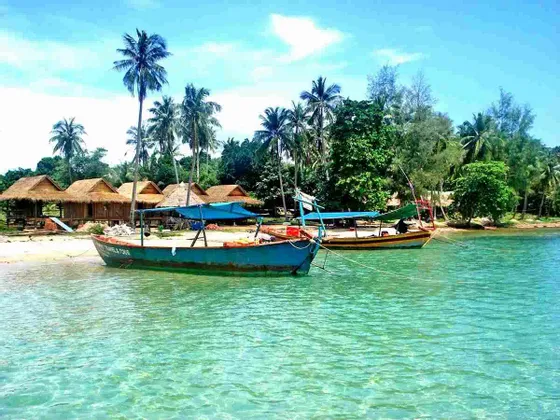 Due barche sono ormeggiate in acque limpide e turchesi, vicino a una spiaggia sabbiosa con capanne di paglia e palme.