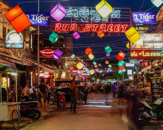 Eine belebte Straße bei Nacht, beleuchtet von einem 'Pub Street' Neonschild und bunten würfelförmigen Laternen, die darüber hängen.