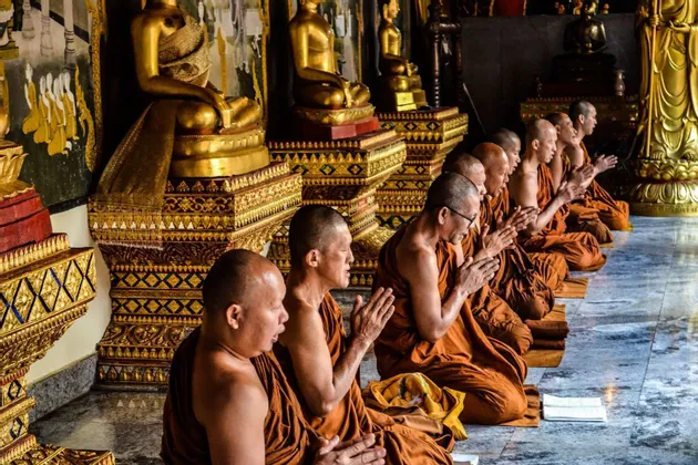 Une rangée de moines bouddhistes en robes oranges agenouillés en prière à l'intérieur d'un temple, avec des statues de Bouddha dorées en arrière-plan.