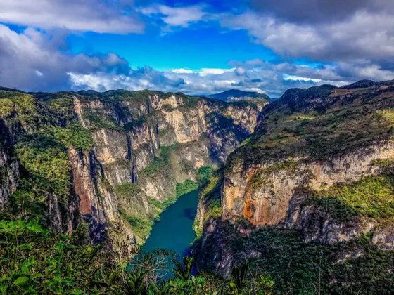 Un río turquesa fluye por un cañón profundo con acantilados empinados y cubiertos de vegetación bajo un cielo azul con nubes dispersas.