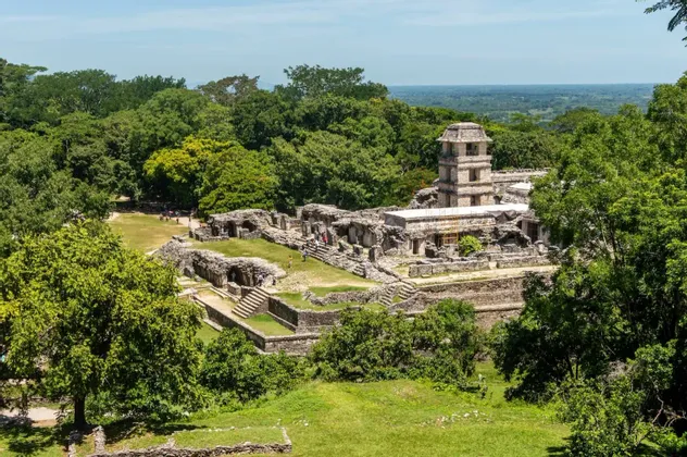 Una vista de gran angular de antiguas ruinas de piedra, incluyendo una torre prominente, rodeada por una densa jungla verde bajo un cielo despejado.