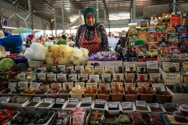 Una donna che indossa un foulard e un grembiule si trova dietro una vivace bancarella del mercato che vende spezie, noodles e altri prodotti alimentari.