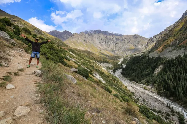 Un uomo con le braccia distese sta su un sentiero di montagna che domina una vasta valle con cime rocciose sotto un cielo parzialmente nuvoloso.