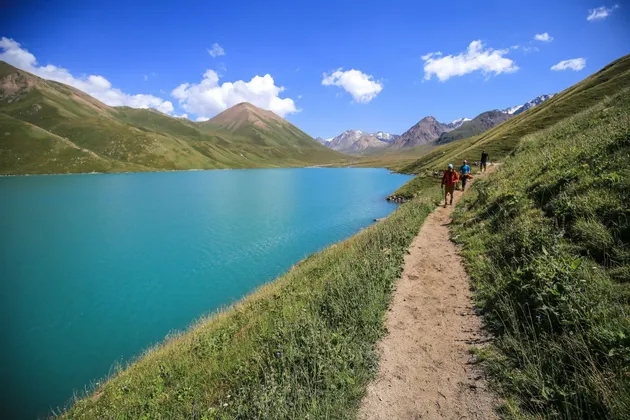 Un viaggio di gruppo WeRoad in trekking su un sentiero sterrato, accanto a un lago alpino turchese, circondato da montagne verdi sotto un cielo azzurro limpido.
