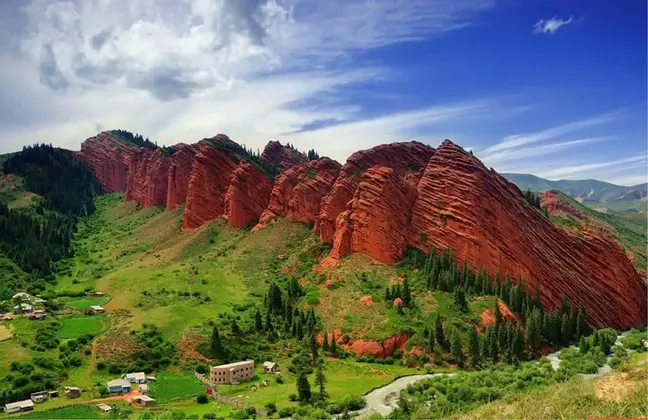 Formazioni rocciose rosse frastagliate dominano una lussureggiante valle verde con un piccolo villaggio e un fiume sotto un cielo parzialmente nuvoloso.