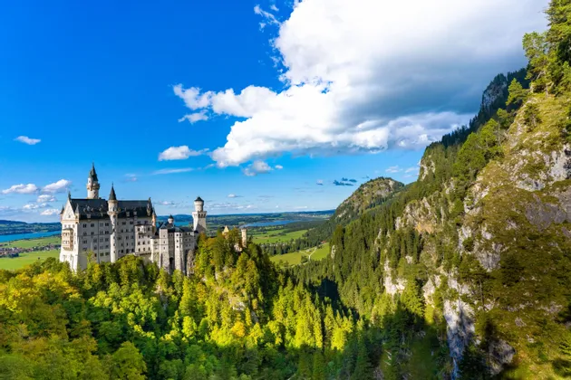 Un grande castello con torri si erge su una collina boscosa, dominando una vasta valle verde e montagne sotto un cielo azzurro con nuvole bianche.
