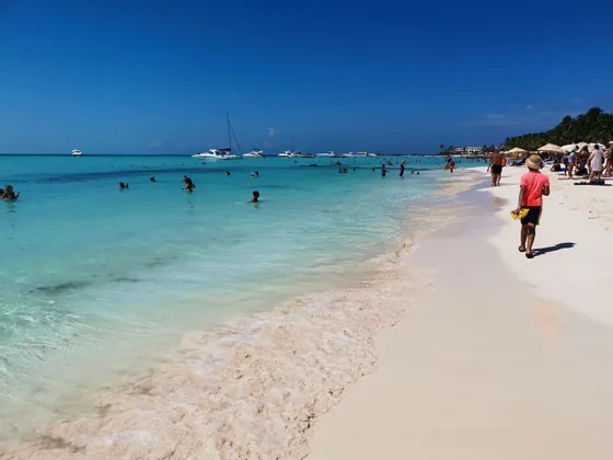 Molte persone si godono una giornata di sole su una spiaggia di sabbia bianca e acqua turchese cristallina, con diverse imbarcazioni all'orizzonte.