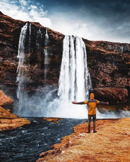 Una persona con una chaqueta amarilla se encuentra en la orilla de un río con los brazos extendidos, frente a una gran cascada que cae por un acantilado rocoso.
