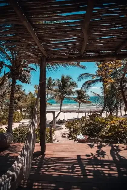 Una vista desde un porche de madera con techo de palos, mirando hacia una playa tropical con palmeras y agua turquesa.