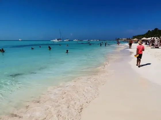 Un día soleado en una playa concurrida donde la gente nada en agua turquesa y camina sobre arena blanca, con barcos visibles en la distancia.